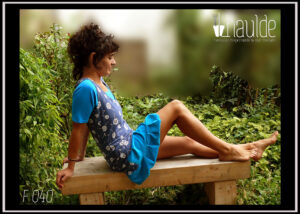 Robe sans manche très échancrée et top manches mi longues en jersey bleu ciel. Robe en jersey bleu jean imprimé pissenlits en graines blanc et volant de velours bleu ciel Vue portée par une femme assise de profil, les jambes sur un banc