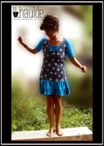 Robe courte sans manche très échancrée et top manches mi longues en jersey bleu ciel. Robe en jersey bleu jean imprimé aigrettes de pissenlit blanches avec en bas deux volants de velours bleu ciel Vue portée par une femme debout sur un banc dans un jardin, de face