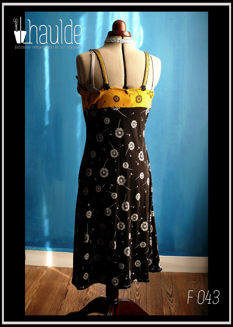 robe mi-longue à bretelles, en jersey jaune imprimé aigrettes de pissenlits noirs jusqu'en dessous du buste et jersey noir imprimé aigrettes de pissenlits blancs pour le bas. Les bretelles sont jaunes piquées de motifs noirs et une dentelle de fleurs noires court en haut de la robe Vue de dos