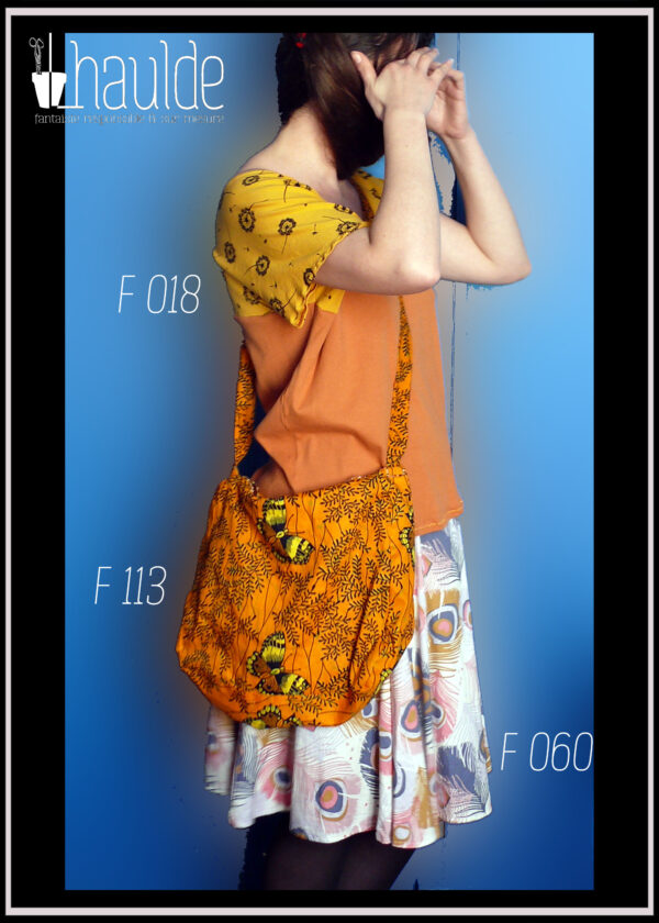 Femme debout de profil portant un tshirt orange à manches courtes jaunes imprimé aigrettes de pissenlit noires, et une jupe à pans amples blanches imprimée plumes de paon bleu jean, rose pâle et ocre