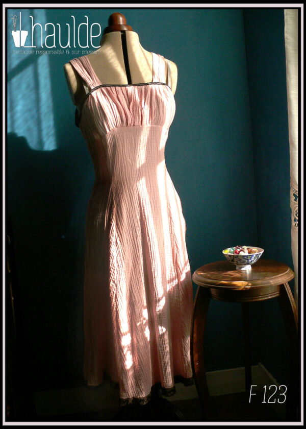 Robe d'été à bretelles en double gaze rose pâle, galon de dentelle noire au bas de la robe, biais noir autour du haut du buste. buste plissé, taille ajustée, jupe évasée. Vue de face au soleil