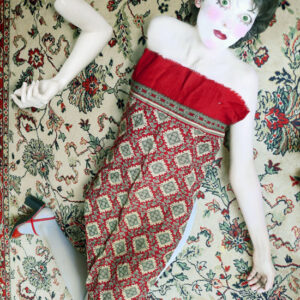 autoportrait déguisée en poupée, yeux et tête agrandis, couverte d'un foulard qui ressemble au tapis sur lequel la doll est posée. Un bras est posé à part à côté