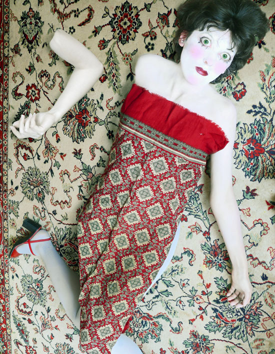 autoportrait déguisée en poupée, yeux et tête agrandis, couverte d'un foulard qui ressemble au tapis sur lequel la doll est posée. Un bras est posé à part à côté
