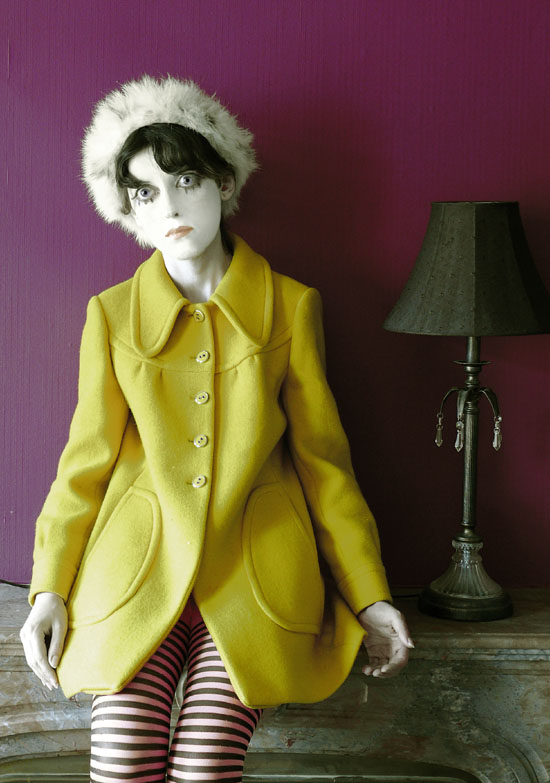 photo couleur, autoportrait déguisée en poupée, yeux et tête agrandis, manteau jaune, posée sur une cheminée devant un mur violet