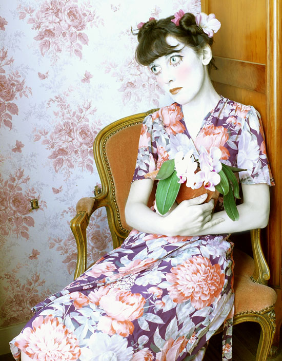 autoportrait déguisée en poupée, yeux et tête agrandis, robe à fleurs, fleurs dans les cheveux, devant une tapisserie à fleurs et avec des orchidées dans les bras