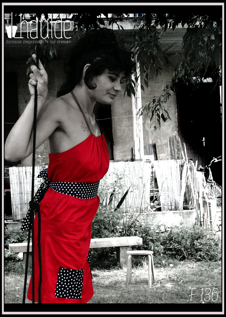 Robe courte dos nu en satin de coton rouge, ceinture et poches plaquées en tissu noir à pois blancs Vue portée de profil par une dame debout sur une balançoire dans un jardin passé en noir et blanc