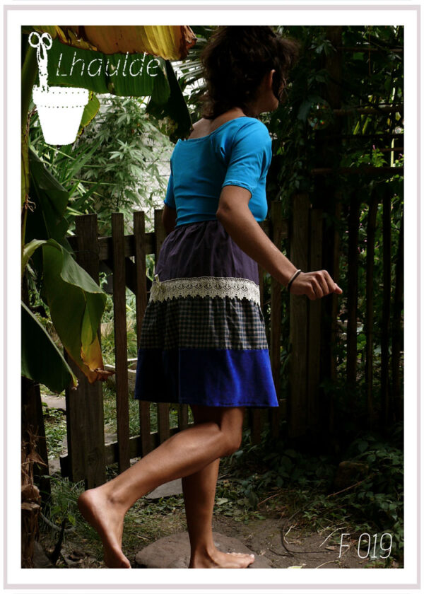 femme debout de dos dans un jardin vêtue d'un tshirt à manches mi longues bleu ciel et de la jupe à pans horizontaux, toile mauve, galon de dentelle, tissu à carreaux vert et gris et le dernier pan en velours bleu roi