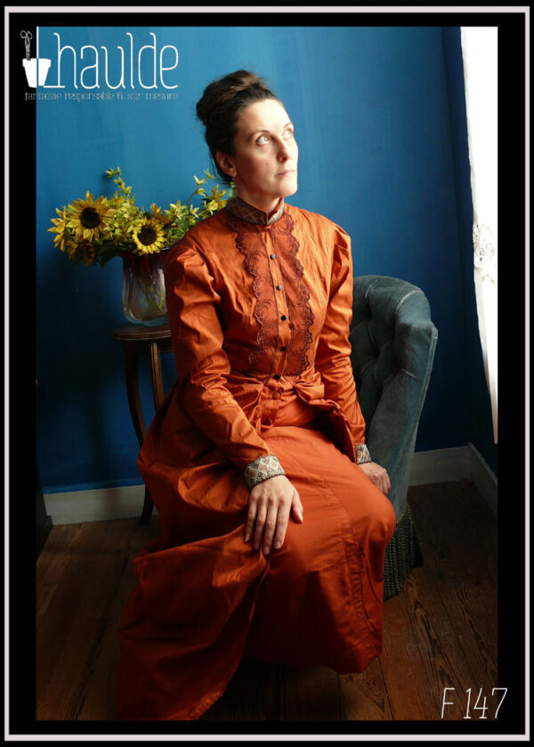 Robe en soie, coupe XIXième, col montant, poitrine ajustée, jupe bouffante sur les reins façon tournure Vue portée sur une femme assise de trois quarts sur un fauteuil