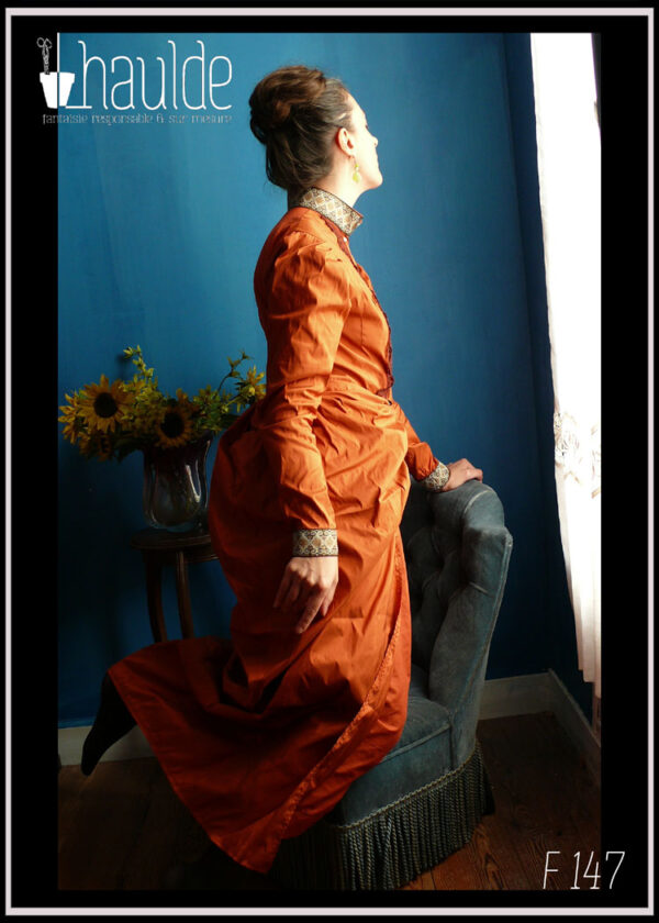 Robe en soie, coupe XIXième, col montant, poitrine ajustée, jupe bouffante sur les reins façon tournure Vue portée sur une femme vue de profil à genoux sur un fauteuil