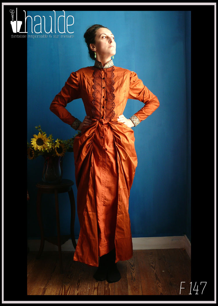 Robe en soie, coupe XIXième, col montant, poitrine ajustée, jupe bouffante sur les reins façon tournure Vue de face portée par une femme debout les mains campées sur les hanches