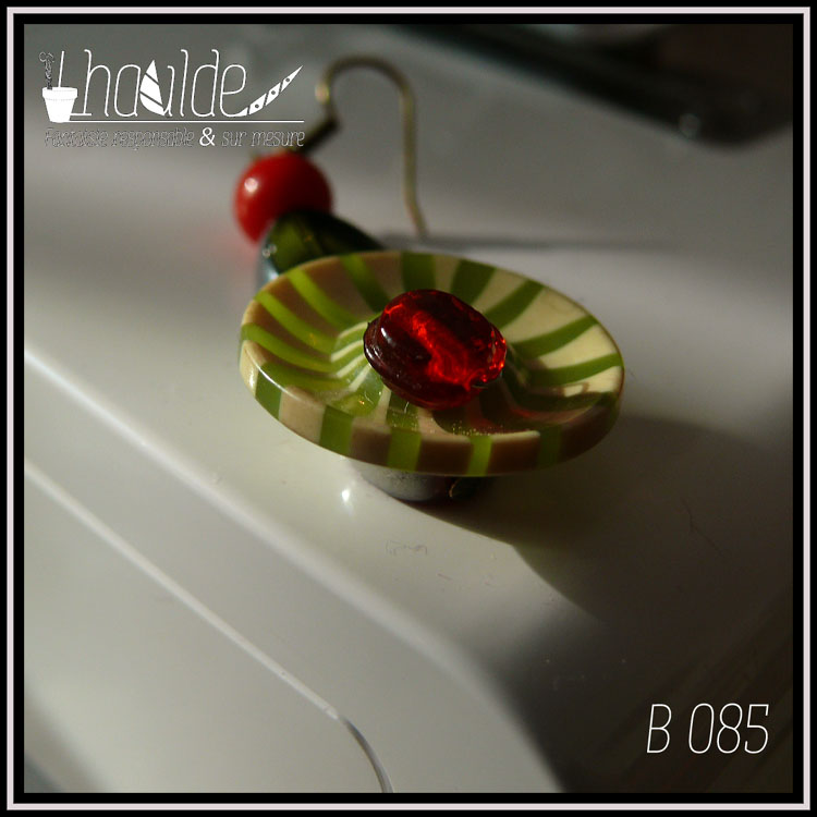 Paire de boucles d'oreilles, crochet acier. Bouton vintage en plastique rayé vert et vert pâle (les rayures partent du centre du bouton vers l'extérieur, comme des rayons) monté sur tige acier avec en son centre une perle en verre rouge avec une tranche en acier, au dos une perle en verre rouge biseauté et au-dessus une perle oblongue en forme de feuille en verre vert avec tranche acier et une perle en verre opaque rouge. Longueur 6,5 cm