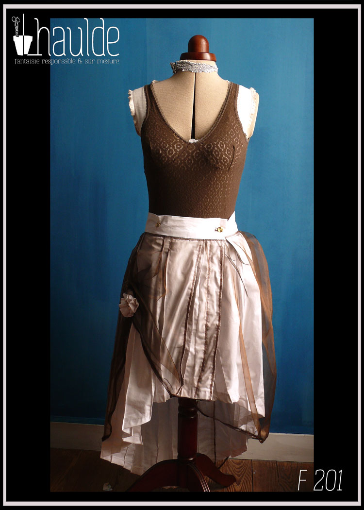 Jupe asymétrique plissée façon kilt en popeline blanche voilée d'organza marron à reflets cuivrés, l'organza est remonté sur le côté droit par une fleur en popeline blanche avec en son centre des boutons et des perles de rocailles dorées et nacrées. Vue de face en pied sur un mannequin couture