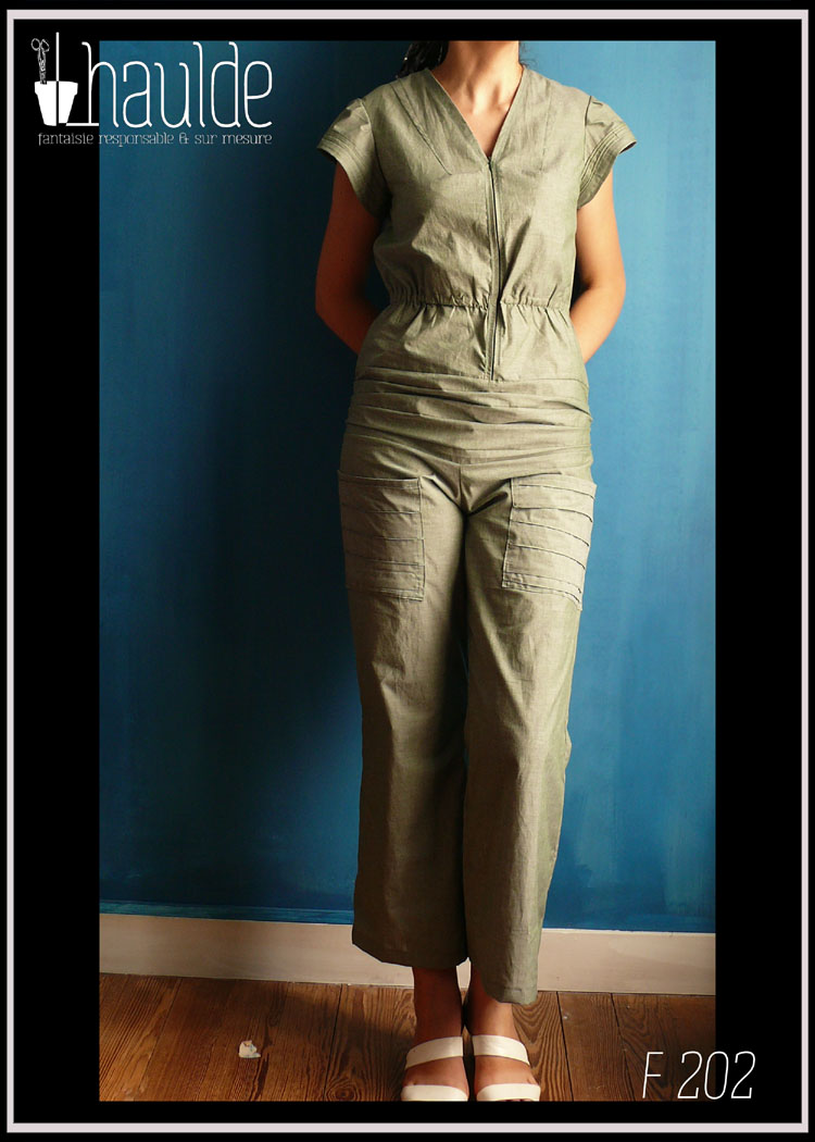 Combinaison manches très courtes et plissé, bande de tissu plissé en bas du ventre et poches plaquées plissées sur le devant des jambes. Zip central devant et taille soulignée par un élastique. Vue portée en pied de face