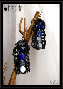 paire de boucles d'oreilles en bande vidéo crochetée montée sur crochet acier avec au centre, alignées à la verticale, quatre perles allant du bleu foncé (en haut) au bleu clair) en bas. Une perle en forme de dodécaèdre en plastique bleu foncé, deux perles ovales plates en verre transparent bleu roi et bleu clair et pour finir une perle plate circulaire en nacre bleu pâle