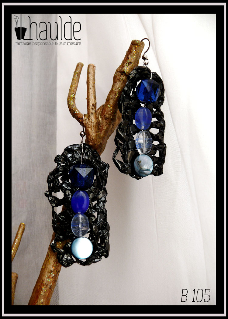 paire de boucles d'oreilles en bande vidéo crochetée montée sur crochet acier avec au centre, alignées à la verticale, quatre perles allant du bleu foncé (en haut) au bleu clair) en bas. Une perle en forme de dodécaèdre en plastique bleu foncé, deux perles ovales plates en verre transparent bleu roi et bleu clair et pour finir une perle plate circulaire en nacre bleu pâle