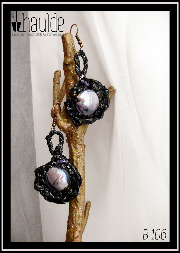 Paire de boucles d'oreilles en bande vidéo crochetée montées sur crochets acier. La bande vidéo est crochetée en rond avec une longue boucle en haut, resserrée par du fil d'acier. Au centre du rond il y a une grosse perle ronde en verre transparent mauve, et quelques perles de rocailles, mauves et cuivrées de ci de là. Vue de la paire de face