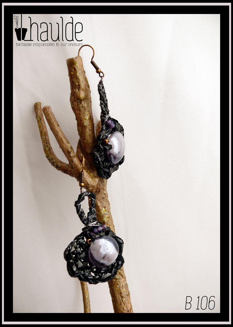 Paire de boucles d'oreilles en bande vidéo crochetée montées sur crochets acier. La bande vidéo est crochetée en rond avec une longue boucle en haut, resserrée par du fil d'acier. Au centre du rond il y a une grosse perle ronde en verre transparent mauve, et quelques perles de rocailles, mauves et cuivrées de ci de là. Vue de la paire de face