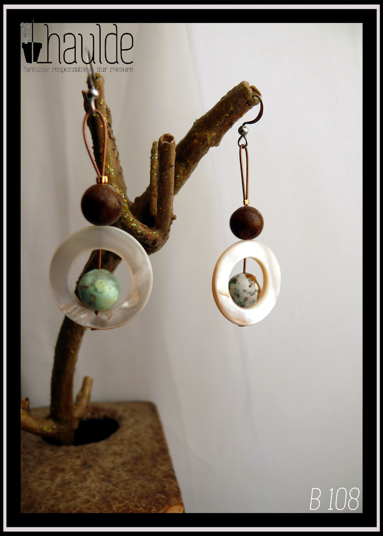 Paire de boucles d'oreilles montées sur crochet. Une perle de dickite marron, un cercle de nacre avec au centre une perle de dickite vert pâle et marron avec des effets de marbrure Elle sont suspendues dans les trous d'une branche d'arbre cirée et pailletée