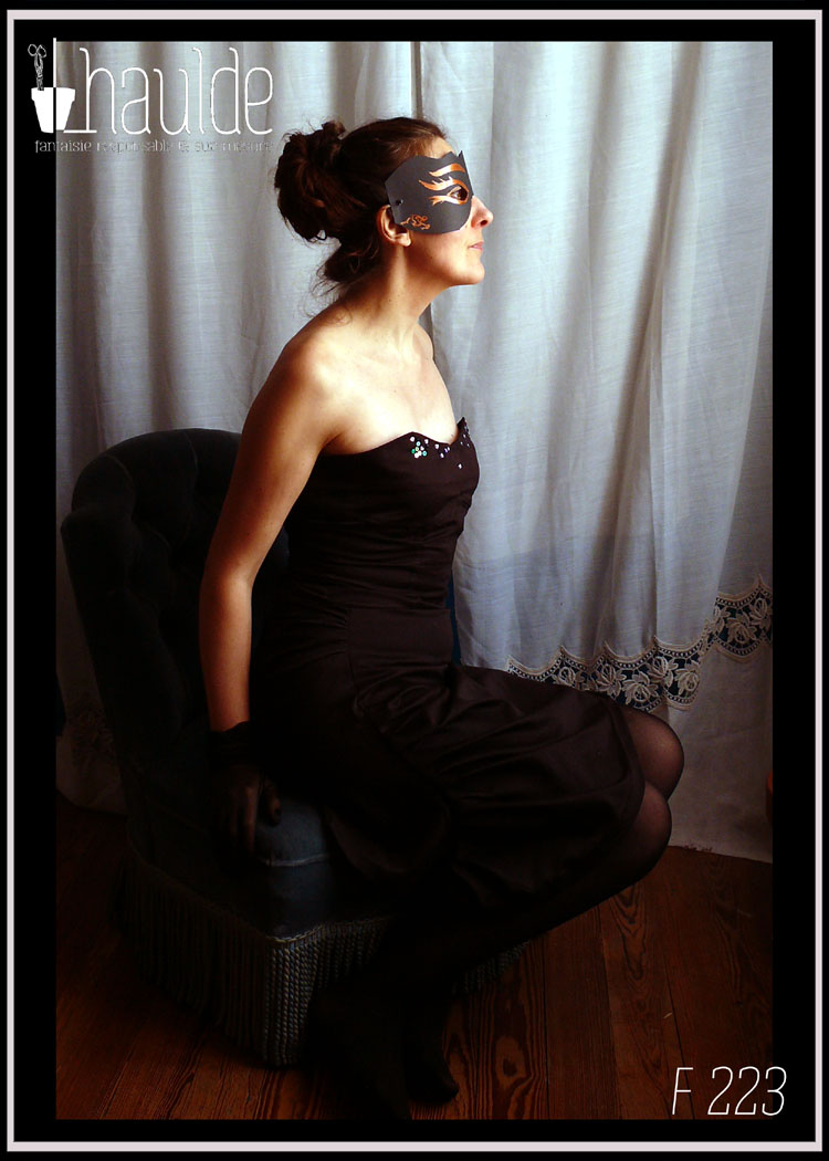 Robe bustier en satin de coton noir courte à jupe asymétrique plissée et haut à pans façon corset. Le haut du bustier est brodé de sequins et de perles de rocailles mauve, violettes, vertes et irisées Vue portée assise