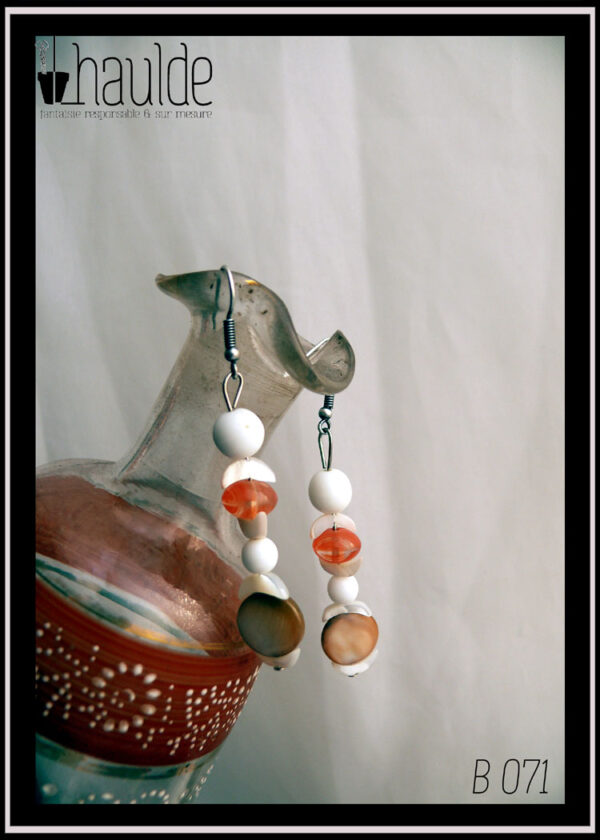 Paire de boucles d'oreilles montées sur crochet acier, perles en nacre blanc et orange et perles en verre blanc et orange Vue sur le rebord d'un vase en verre