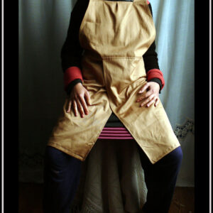 femme assise portant le tablier en question, haut sur la poitrine, grosses bretelles croisées dans le dos, pans fendus devant et derrière
