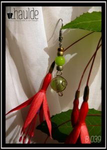 Boucles montées sur crochet, perles en verre alignées à la verticale, deux grosses rocailles dorées, une perle ronde verte, une grosse rocaille dorée, une perle verte transparente entourée de boucles d'acier Vue entourée de fleurs de fuchsia