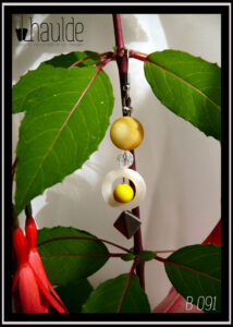 Paire de boucles d'oreilles. Crochet acier. Perles en nacre, verre et acier montées sur tige acier. Perle plate en nacre jaune, perle plate creuse circulaire en nacre blanc avec au centre une perle en verre opaque jaune et pour finir une perle carrée biseautée en acier. Longueur 6,5 cm Vue suspendue à une branche de fuchsia