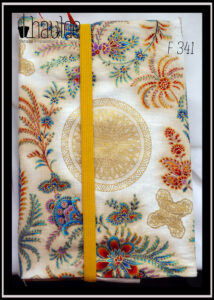 Protège livre en tissu écru imprimé motifs végétaux et floraux d'esthétique asiatique multicolores et bordés de doré. Vu fermé par un élastique jaune