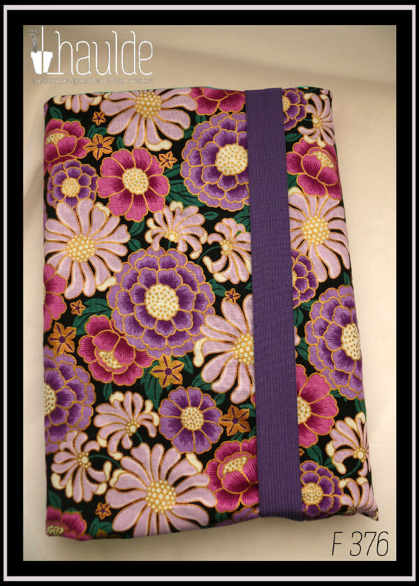 Protège livre en tissu noir imprimé grosses fleurs mauves, violettes et roses, avec du doré, vu fermé par un gros élastique violet