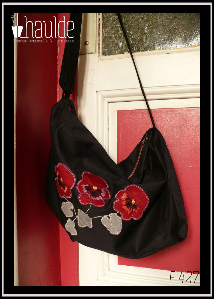 Sac en sergé noir, grand format horizontal à bandoulière, avec des appliqués en forme de pensées rouges avec leurs feuilles. Le centre des fleurs, les contours et les tiges sont brodés à la machine Vu suspendu à une poignée de porte