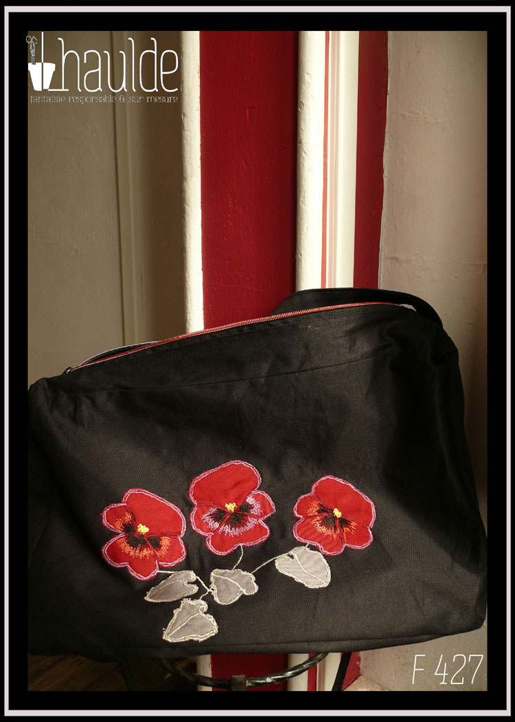 Sac en sergé noir, grand format horizontal à bandoulière, avec des appliqués en forme de pensées rouges avec leurs feuilles. Le centre des fleurs, les contours et les tiges sont brodés à la machine Vu suspendu à une poignée de porte