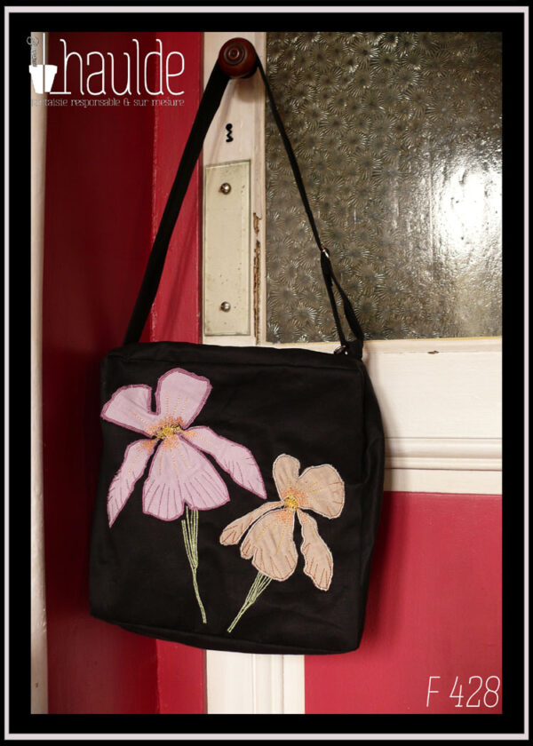 sac en sergé noir format carré à bandoulière avec des appliqués mauves et beiges en forme d'iris, le centre des fleurs et leurs pétales sont brodés à la machine ainsi que les tiges Vu suspendu à une poignée de porte