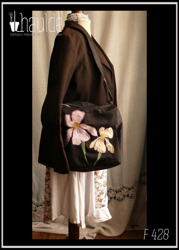 sac en sergé noir format carré à bandoulière avec des appliqués mauves et beiges en forme d'iris, le centre des fleurs et leurs pétales sont brodés à la machine ainsi que les tiges Vu porté sur un mannequin de couture