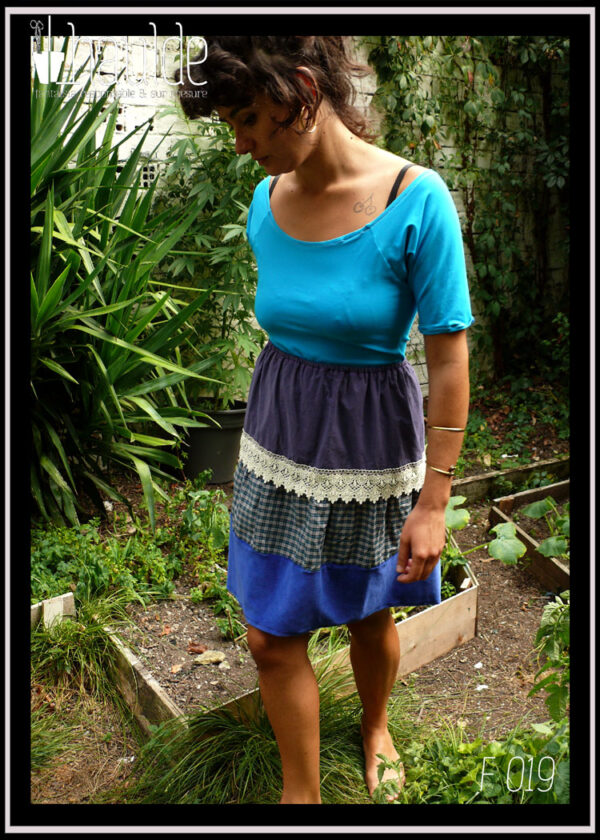 femme debout dans un jardin vêtue d'un tshirt à manches mi longues bleu ciel et de la jupe à pans horizontaux, toile mauve, galon de dentelle, tissu à carreaux vert et gris et le dernier pan en velours bleu roi