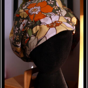 Casquette bouffante en toile enduite imprimée de grosses fleurs oranges jaunes et blanches sur fond vert foncé, le graphisme est une version modernisée de designs des années 70 Vu sur une tête de mannequin, de face