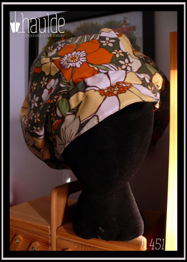 Casquette bouffante en toile enduite imprimée de grosses fleurs oranges jaunes et blanches sur fond vert foncé, le graphisme est une version modernisée de designs des années 70 Vu sur une tête de mannequin, de face