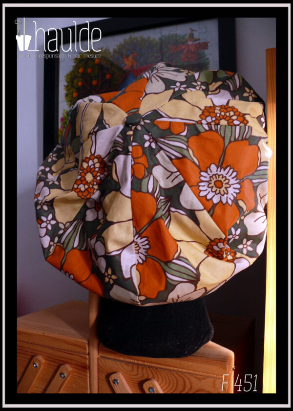 Casquette bouffante en toile enduite imprimée de grosses fleurs oranges jaunes et blanches sur fond vert foncé, le graphisme est une version modernisée de designs des années 70 Vu sur une tête de mannequin, de dos, on voit les huit pans qui forment la casquette et le bouton recouvert du même tissu au centre