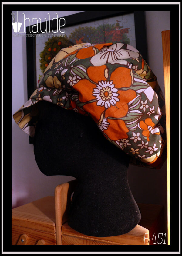 Casquette bouffante en toile enduite imprimée de grosses fleurs oranges jaunes et blanches sur fond vert foncé, le graphisme est une version modernisée de designs des années 70 Vu sur une tête de mannequin, de profil