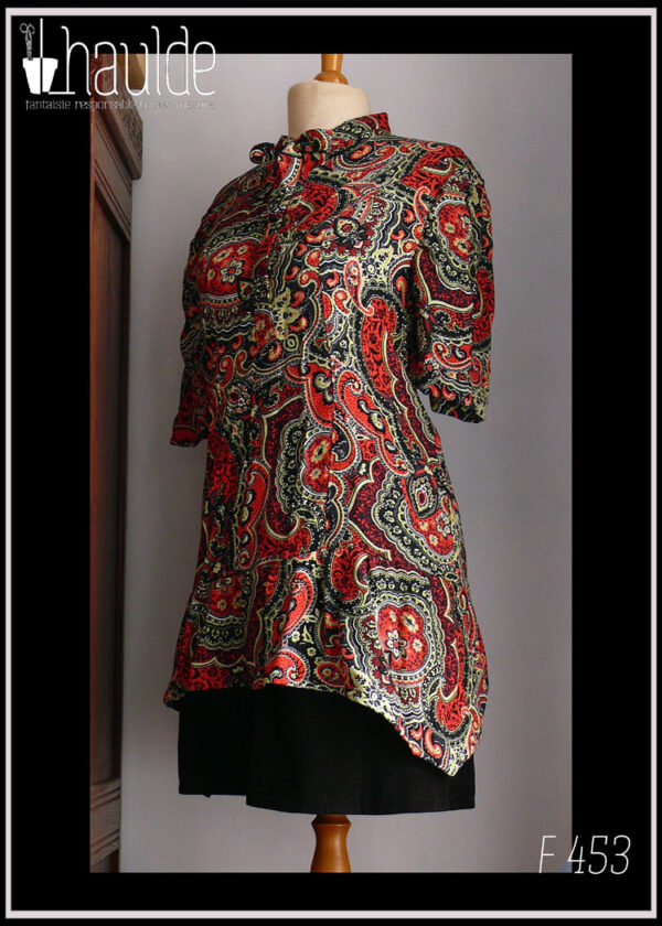 Tunique col lavallière et polo, manches trois quarts, asymétrique, plus longue côté gauche (à mi cuisse environ) en viscose imprimée motifs paisley vert clair et rouge vif sur fond noir. Vu sur un mannequin de couture avec une mini-jupe noire