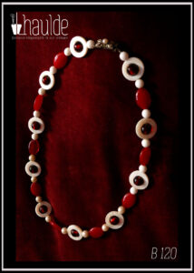 Collier composé de perles en verre blanc opaque alternées avec des cercles de nacre ayant en leur centre une perle en verre rouge à coeur métallique, et des perles oblongues en verre rouge opalescent. Au centre les perles blanches sont remplacées par des fausses perles de nacre Le fermoir vintage est décoré d'une fausse petite perle et se pince pour s'ouvrir Vu de dessus sur du velours rouge bordeaux