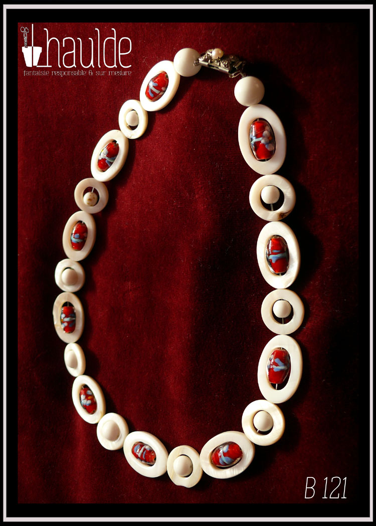 Collier constitués de perles en verre blanc opaque alterné avec des ellipses de nacre ayant au centre une perle oblongue de verre de bohème rouge à inclusion fleurie bleue, blanche et jaune, cercles de nacre avec au centre une perle blanche vu de dessus posé sur du velours rouge bordeaux Le fermoir vintage est décoré d'une fausse petite perle et se pince pour s'ouvrir