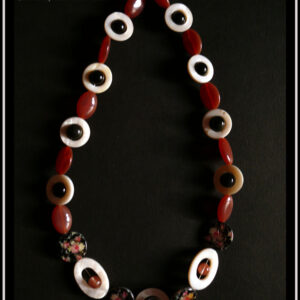 Collier en cercles de nacre alternés avec des perles en verre oblongues couleur vieux rose foncé. Au centre devant il y a une perle en verre de bohème noire à inclusion en forme de fleur bleue et rose, au centre d'un cercle de nacre plus grand que les autres/ De chaque côté sont des perles plates nacrées noir avec des roses imprimées et des ellipses de nacre avec une perle en cristal rose au centre. Les autres cercles de nacre on une perle en verre noir en leur centre Vu posé sur une feuille de papier noir