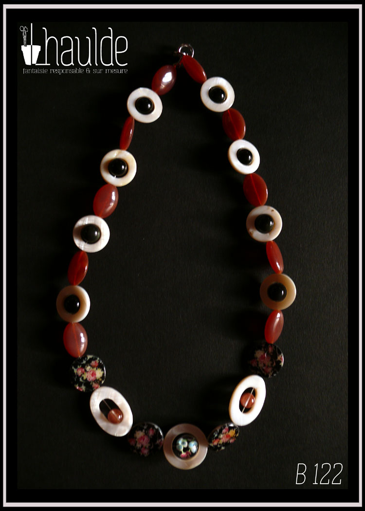 Collier en cercles de nacre alternés avec des perles en verre oblongues couleur vieux rose foncé. Au centre devant il y a une perle en verre de bohème noire à inclusion en forme de fleur bleue et rose, au centre d'un cercle de nacre plus grand que les autres/ De chaque côté sont des perles plates nacrées noir avec des roses imprimées et des ellipses de nacre avec une perle en cristal rose au centre. Les autres cercles de nacre on une perle en verre noir en leur centre Vu posé sur une feuille de papier noir