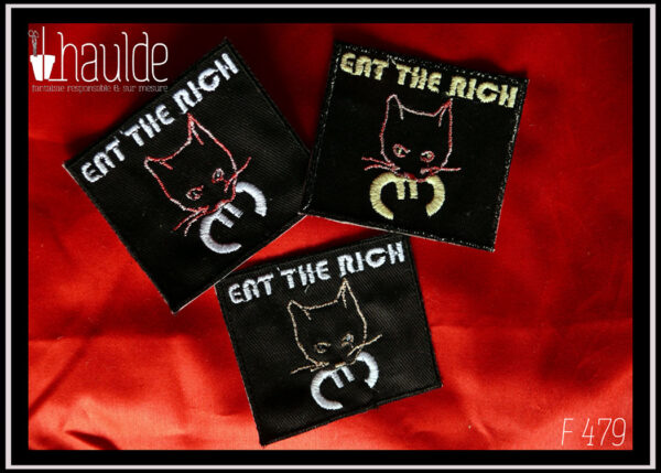 trois patchs "eat the rich" (mange les riches) presque carrés noirs, une tête de chat qui tient dans sa gueule le symbole de l'euro, le texte "eat the rich" est tout en haut du patch. La tête du chat est une simple ligne avec les yeux et la moustache, soit rose chamarré soit rose pâle, le texte et le symbole euro sont soit en blanc soit légèrement jaune (fil phosphorescent) Vu sur un morceau de tissu rouge