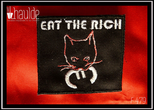 le patch "eat the rich" (mange les riches) tête de chat tenant le symbole euro dans sa gueule avec le texte au-dessus, sur fond noir) version fil blanc (pour le texte, les pupilles et le symbole euro, la tête du chat est dessinée par une ligne rose chamarrée