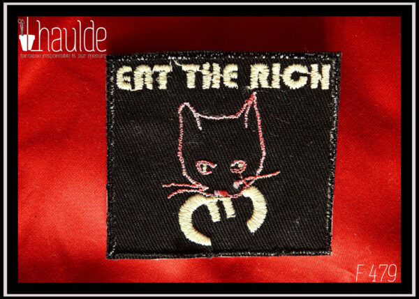 le patch "eat the rich" (mange les riches) tête de chat tenant le symbole euro dans sa gueule avec le texte au-dessus, sur fond noir) version fil phosphorescent (pour le texte, les pupilles et le symbole euro, la tête du chat est dessinée par une ligne rose chamarrée