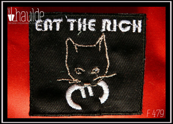 le patch "eat the rich" (mange les riches) tête de chat tenant le symbole euro dans sa gueule avec le texte au-dessus, sur fond noir) version fil blanc (pour le texte, les pupilles et le symbole euro, la tête du chat est dessinée par une ligne rose pâle