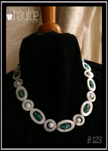 Collier constitué d'ellipses de nacre blanc alternées avec des cercles de nacre blanc, à l'intérieur de chaque ellipse il y a une perle en verre de Bohème oblongue vert pâle à incrustation fleurs turquoise et rouge. A l'intérieur de chaque cercle une perle en verre blanc opaque. Le fermoir est un mousqueton flanqué de deux grosses perles en verre blanc opaque et de cercle de nacre avec une petite perle turquoise au centre Vu autour du cou d'un mannequin de couture vêtu de velours noir