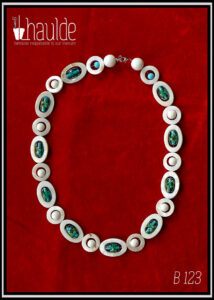 Collier constitué d'ellipses de nacre blanc alternées avec des cercles de nacre blanc, à l'intérieur de chaque ellipse il y a une perle en verre de Bohème oblongue vert pâle à incrustation fleurs turquoise et rouge. A l'intérieur de chaque cercle une perle en verre blanc opaque. Le fermoir est un mousqueton flanqué de deux grosses perles en verre blanc opaque et de cercle de nacre avec une petite perle turquoise au centre Vu de dessus posé sur du velours rouge vif