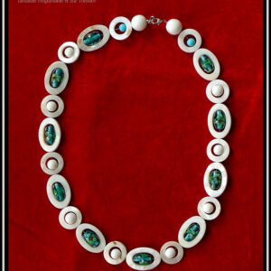 Collier constitué d'ellipses de nacre blanc alternées avec des cercles de nacre blanc, à l'intérieur de chaque ellipse il y a une perle en verre de Bohème oblongue vert pâle à incrustation fleurs turquoise et rouge. A l'intérieur de chaque cercle une perle en verre blanc opaque. Le fermoir est un mousqueton flanqué de deux grosses perles en verre blanc opaque et de cercle de nacre avec une petite perle turquoise au centre Vu de dessus posé sur du velours rouge vif